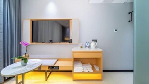 A modern hotel room with a large flat-screen TV mounted on the wall, a wooden vanity with a mirror, a small table with a vase of flowers, and a shelf with towels and toiletries. - Guangyi Hotel (Quanzhou)