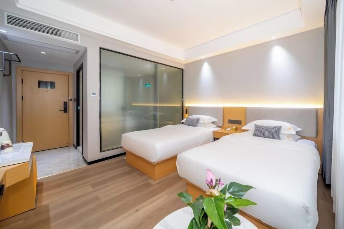 A hotel room with two beds, a desk, a chair, a plant, and a sliding glass door.