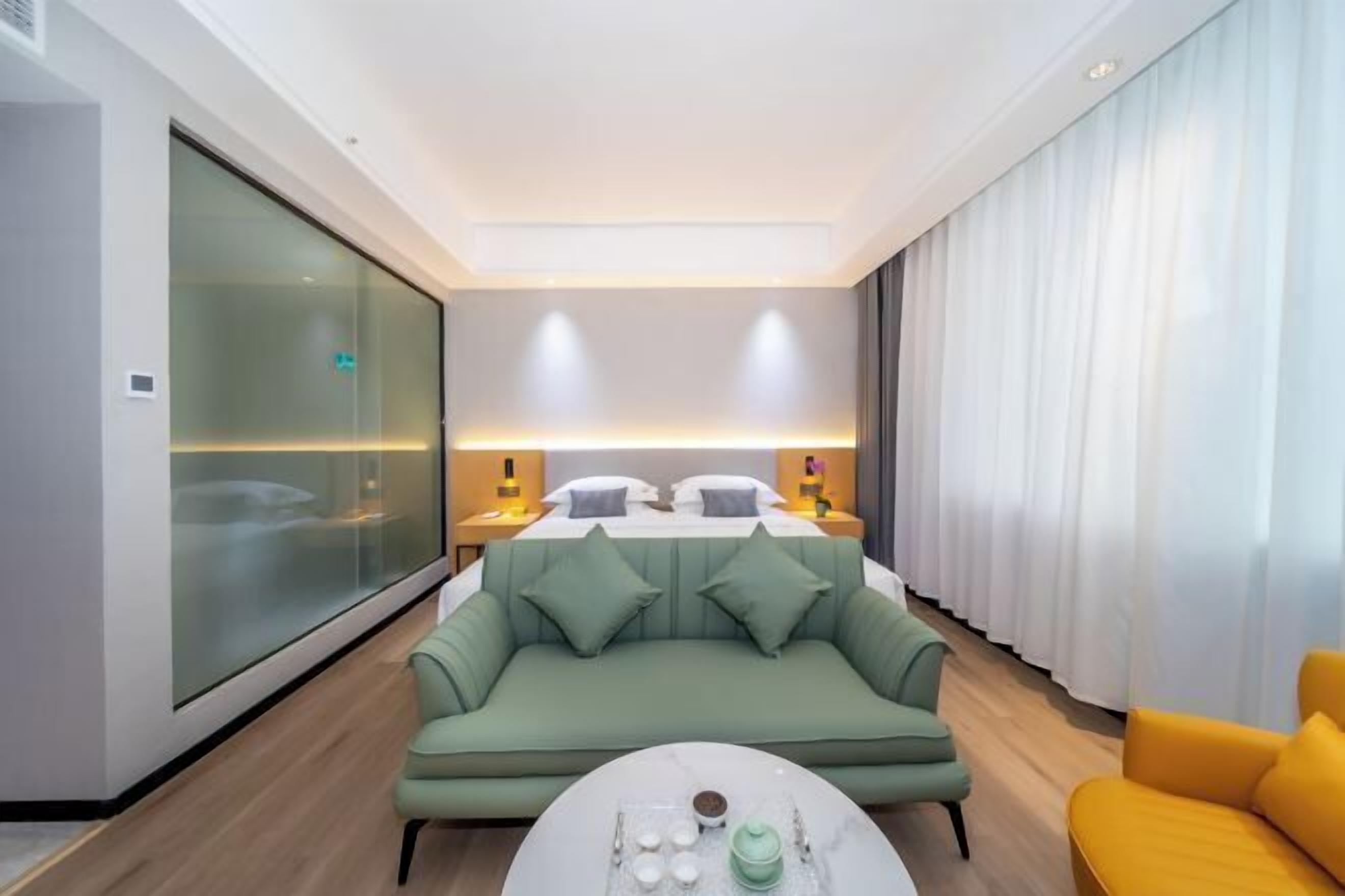 A modern hotel room with a bed, a green sofa, a yellow armchair, and a round coffee table.