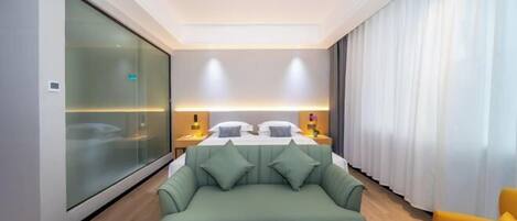 A modern hotel room with a bed, a green sofa, a yellow armchair, and a round coffee table.