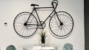 Un comedor con una mesa de vidrio y dos sillas, una bicicleta colgada en la pared, y una pequeña planta en maceta.