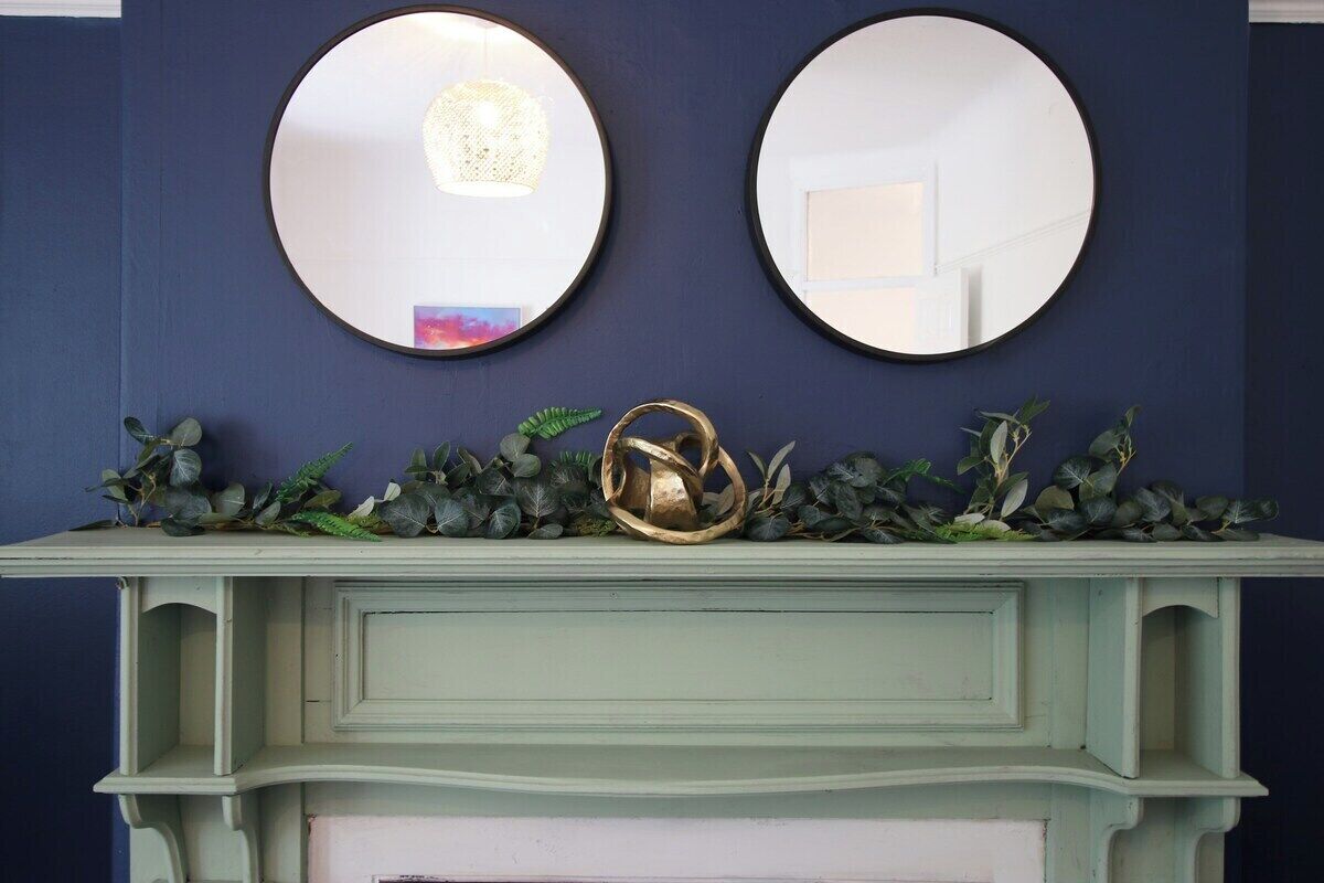 A blue wall with two round mirrors and a white mantelpiece with a decorative metal object and greenery.