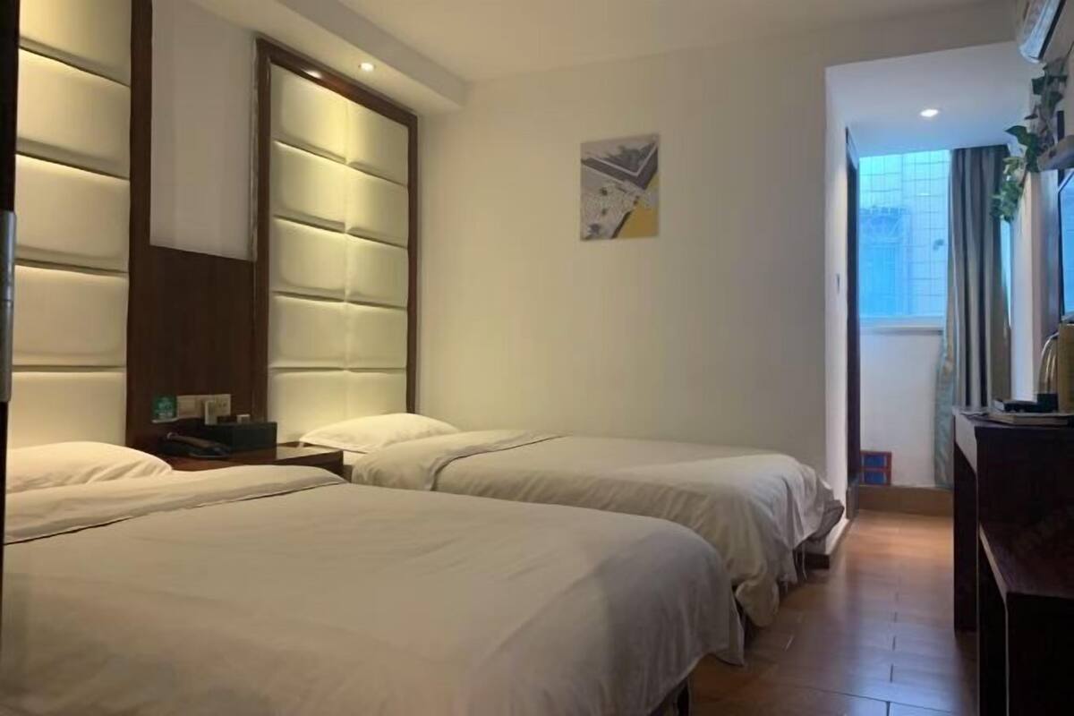A hotel room with two beds, a large window with curtains, a wall-mounted artwork, and a wooden headboard.