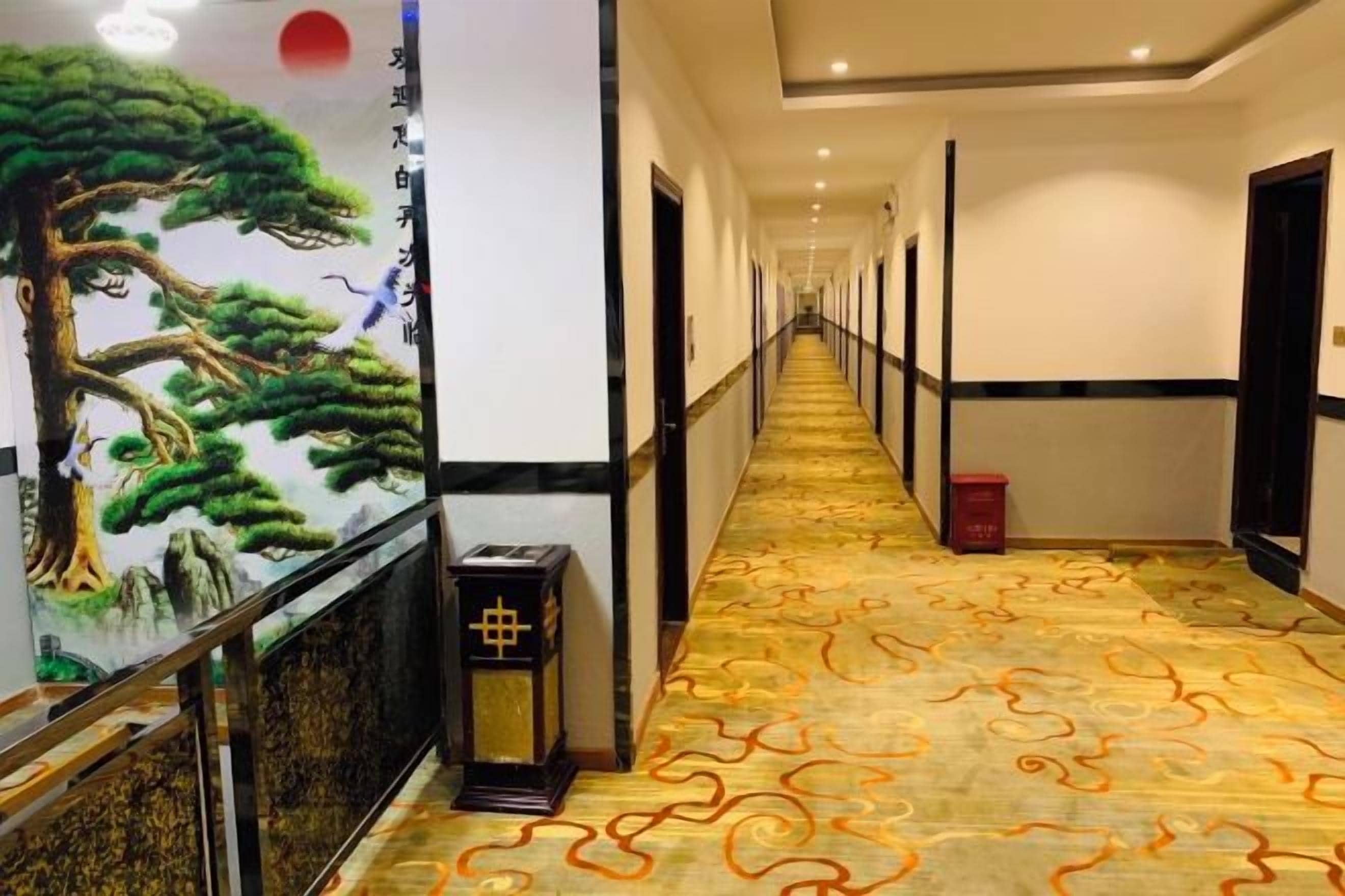 A hotel corridor with a patterned carpet, a traditional mural, and a red trash bin.