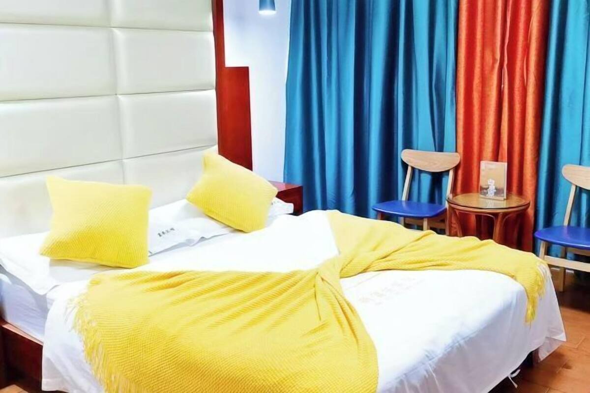 A hotel room with a bed, yellow pillows, a yellow blanket, a wooden nightstand, a small table with a book, and blue curtains.