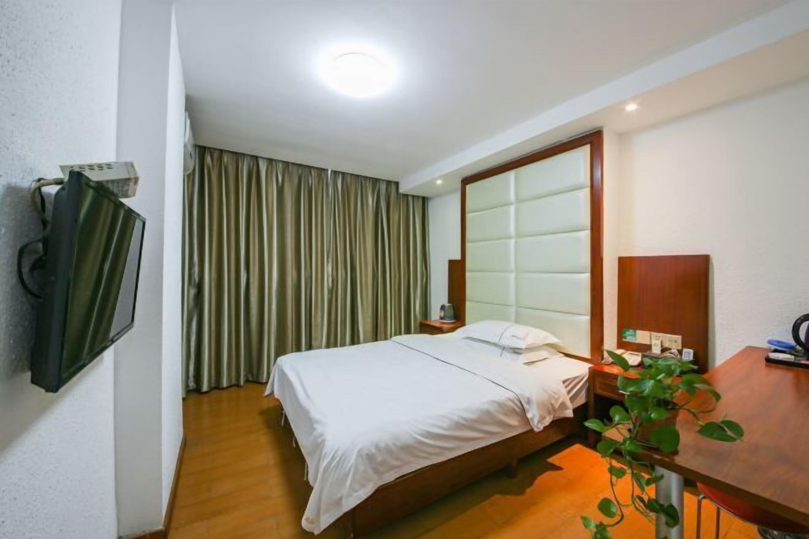 Ein Hotelzimmer mit Bett, Nachttisch, Spiegel, Pflanze und einem fenster mit Vorhängen.
