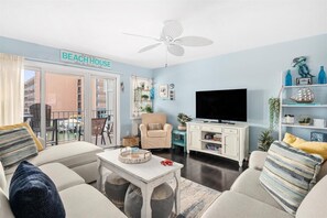A living room with a sofa, armchairs, a coffee table, a TV, and a ceiling fan. There is a sign on the wall that reads "BEACH HOUSE".
