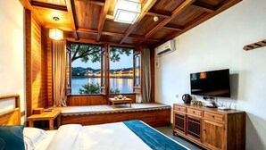 Een slaapkamer met een groot bed, houten plafond, een televisie, een raam met uitzicht op een wateroppervlak en een zithoek bij het raam.