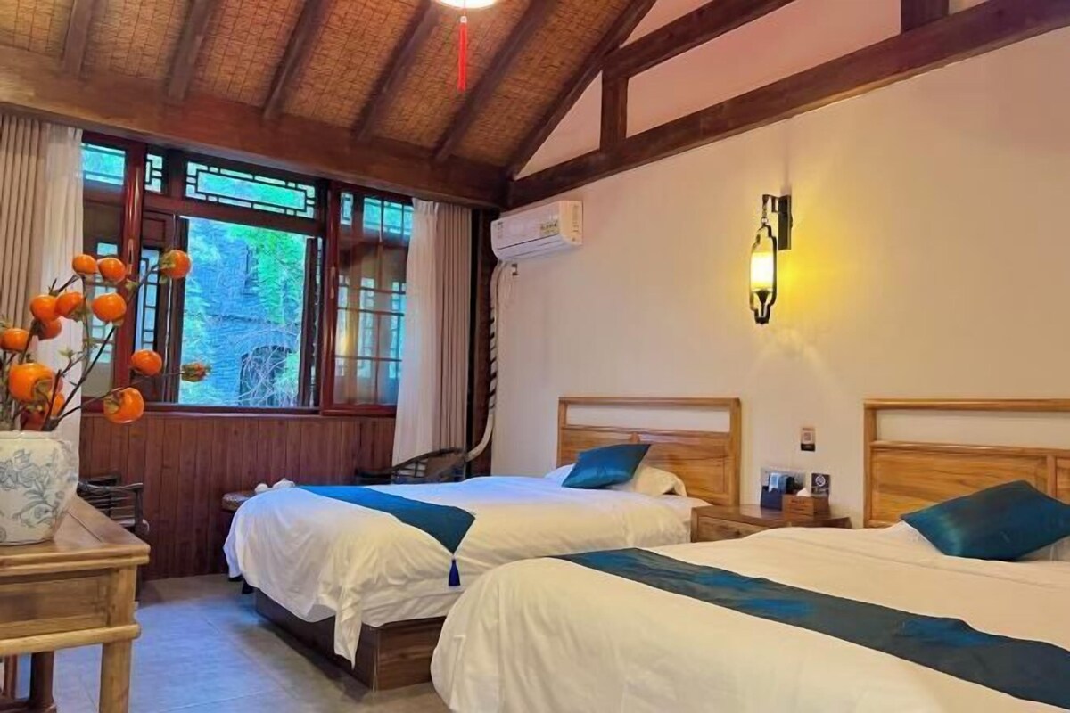 Een hotelkamer met twee bedden, houten hoofdborden, nachtkastjes, een vaas met oranje bloemen en een raam met gordijnen.