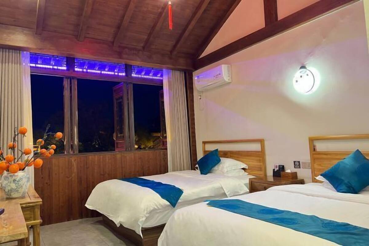 Een hotelkamer met twee bedden, houten balken in het plafond, een airconditioning, nachtkastje en een raam met gordijnen.