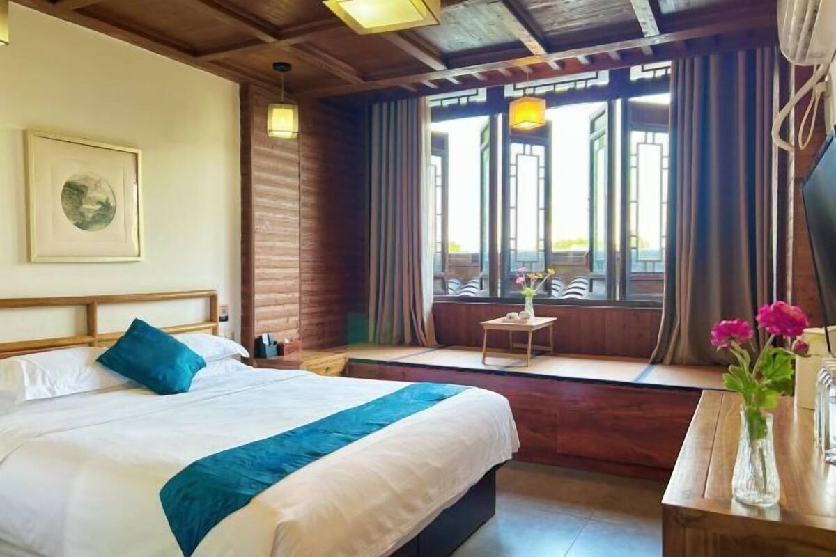 Een hotelkamer met een groot bed, een klein tafeltje met een bloemenvaas, en een raam met gordijnen.