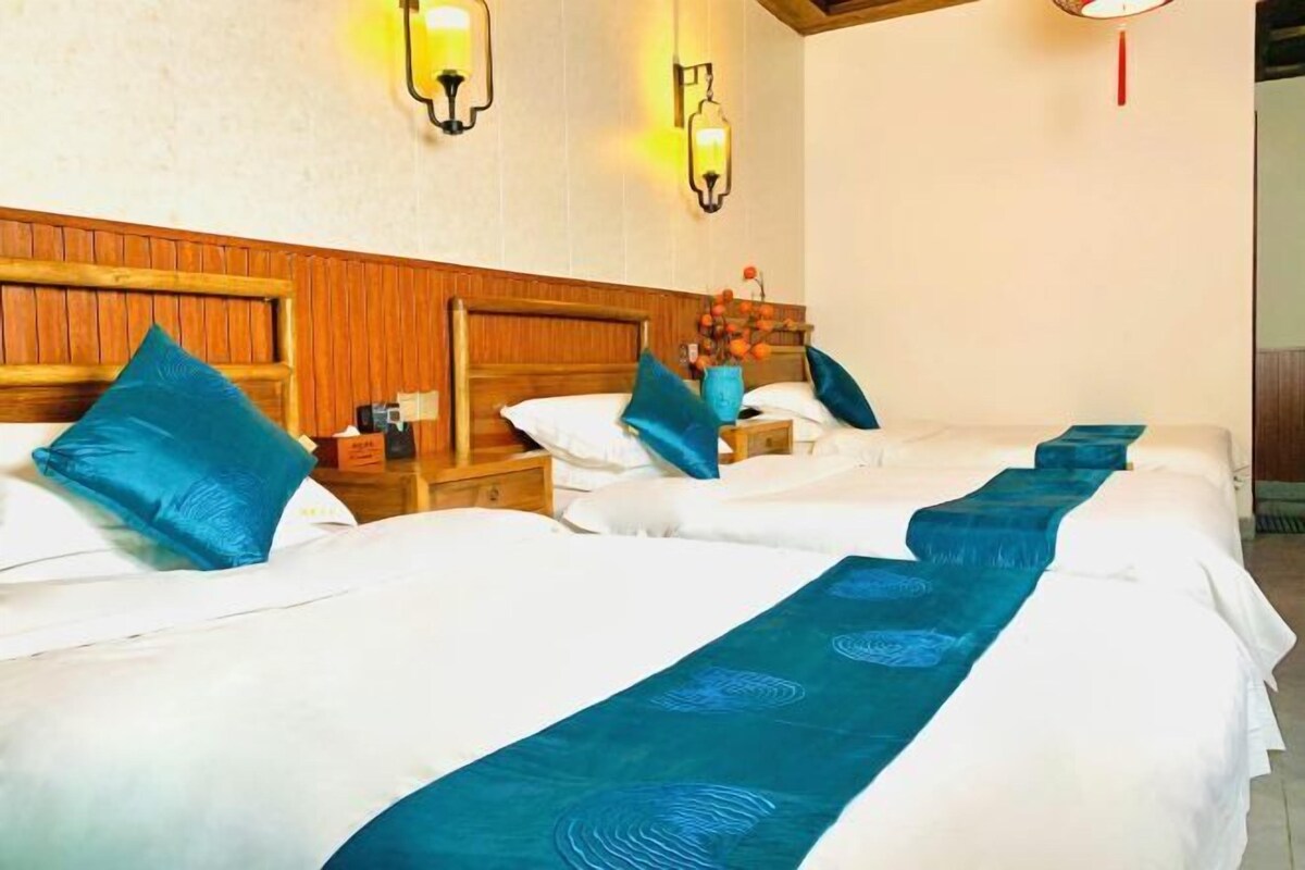 Een hotelkamer met drie bedden, elk met een blauw kleed en witte beddengoed. Er zijn wandlampen en een houten hoofdbord.