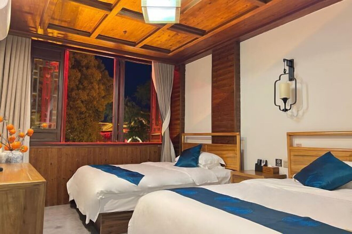 Een hotelkamer met twee bedden, houten hoofdeinden, een nachtkastje, een lamp en een raam met gordijnen.
