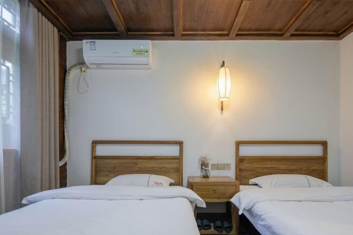 Een hotelkamer met twee bedden, houten hoofdeinden, een wandairconditioner, een bedlampje en een klein nachtkastje met een vaas.