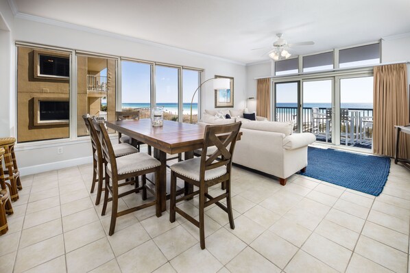 A spacious living area with a dining table, chairs, and a sofa. Large windows offer a view of the beach.