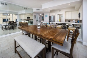 A spacious dining area with a large wooden table and cushioned benches, a kitchen with modern appliances, and large windows offering a view of the outside.