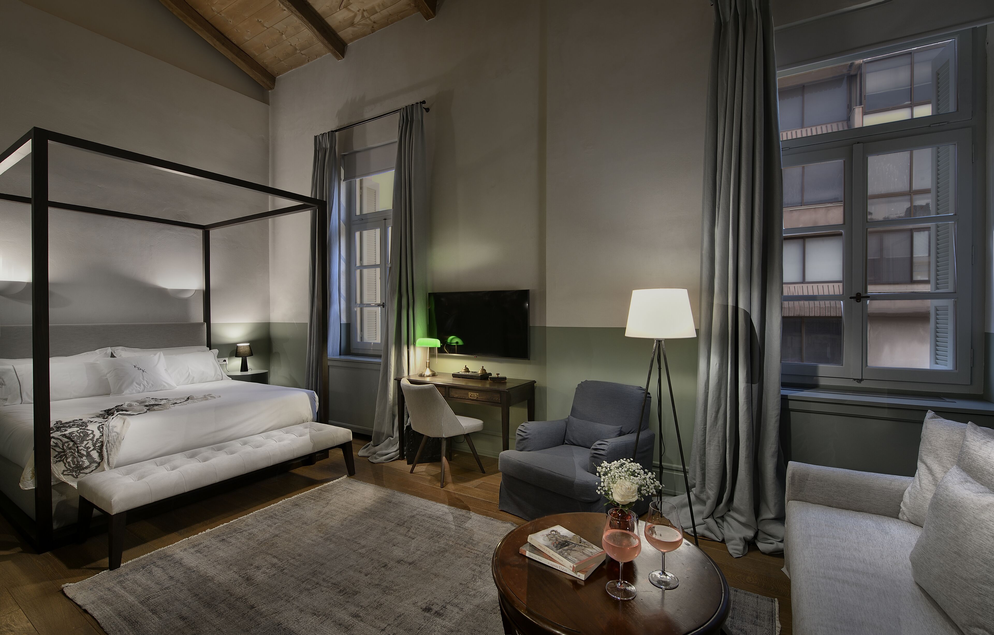 Hotelkamer met een hemelbed, een bureau met een computer, een grijze fauteuil, een bank en een salontafel met een boek en glazen.