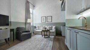 A compact living space with a sofa, a small round table, a TV mounted on the wall, a kitchen area with a sink, and a window with curtains. - Athinaidos Avant Garde Loft by K&K (Αθήνα)