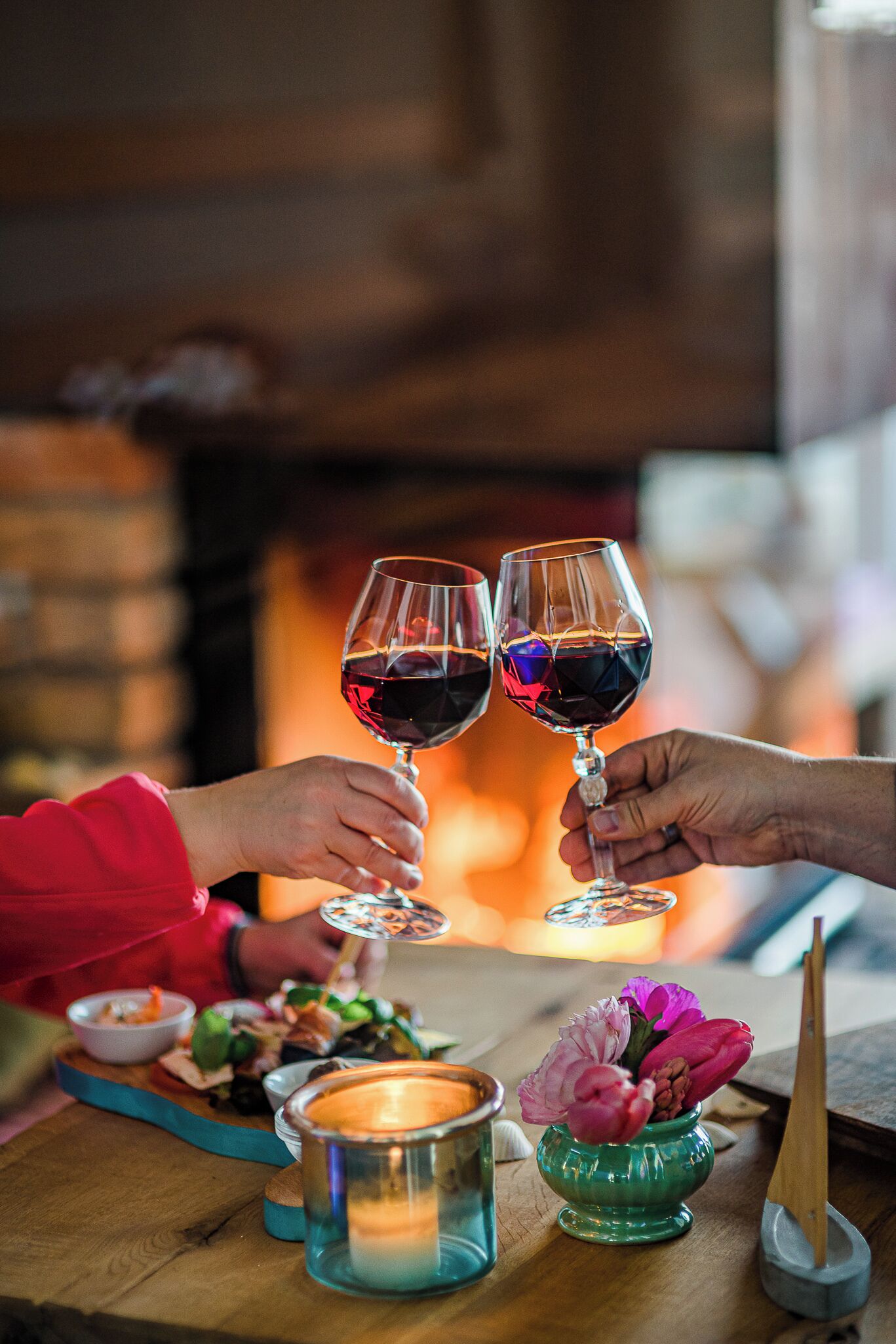 Dos personas brindando con copas de vino, una vela y un pequeño florero con flores sobre una mesa de madera.