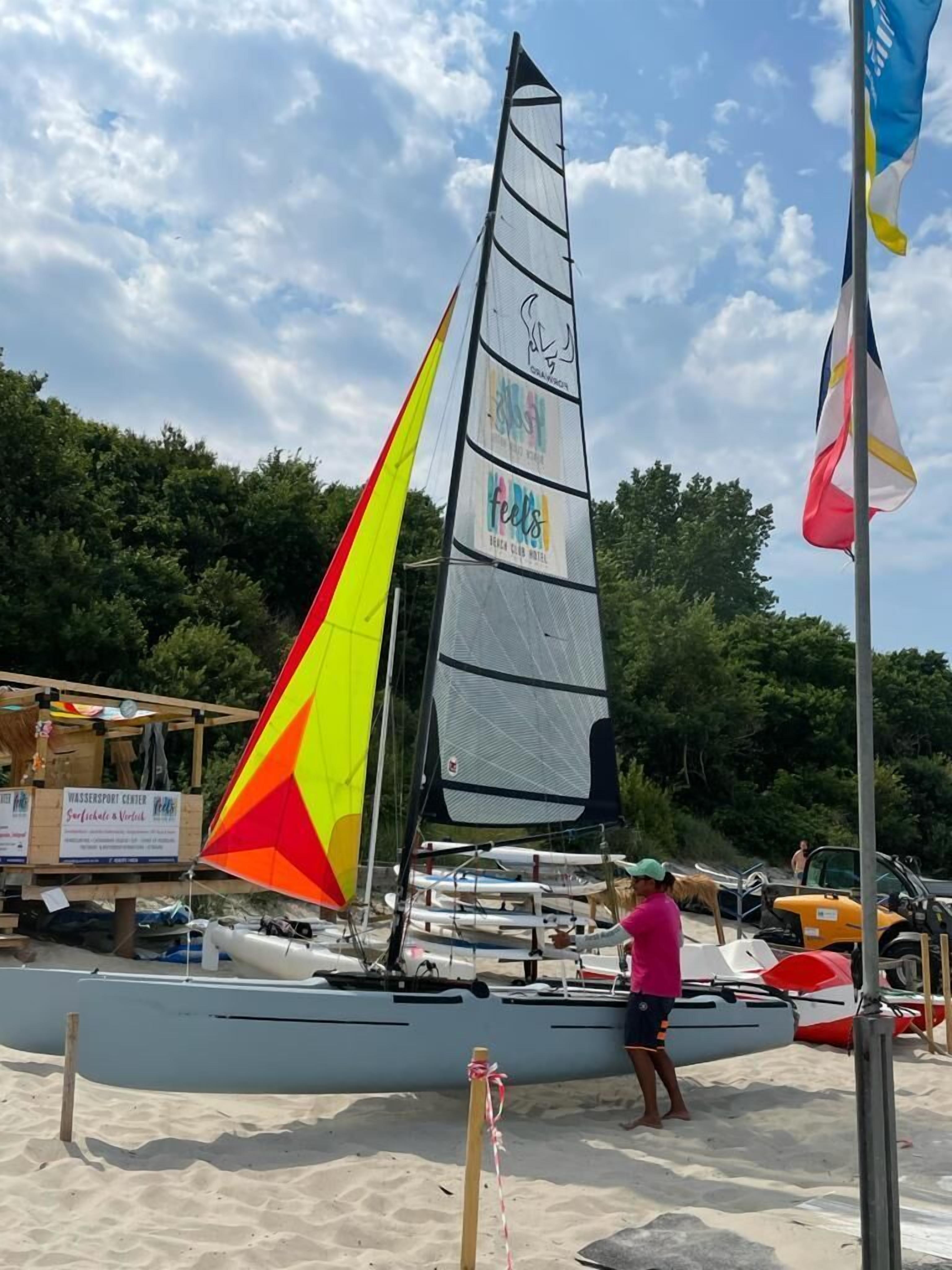 A sailboat with a colorful sail docked on a sandy beach, a signboard in the background, and a flagpole with a flag.
