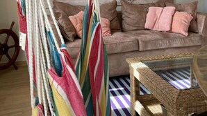 Een woonkamer in strandthema met een hangmat, een bank met kussens, een rieten tafeltje en een wanddecoratie van een strand.