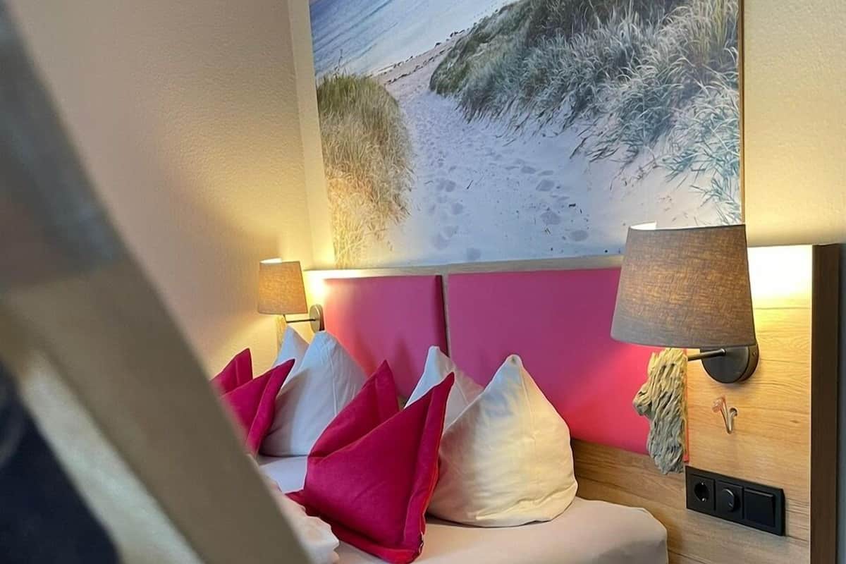 Een slaapkamer met een bed, kussens, een nachtkastje, een lamp en een schilderij van een strand.