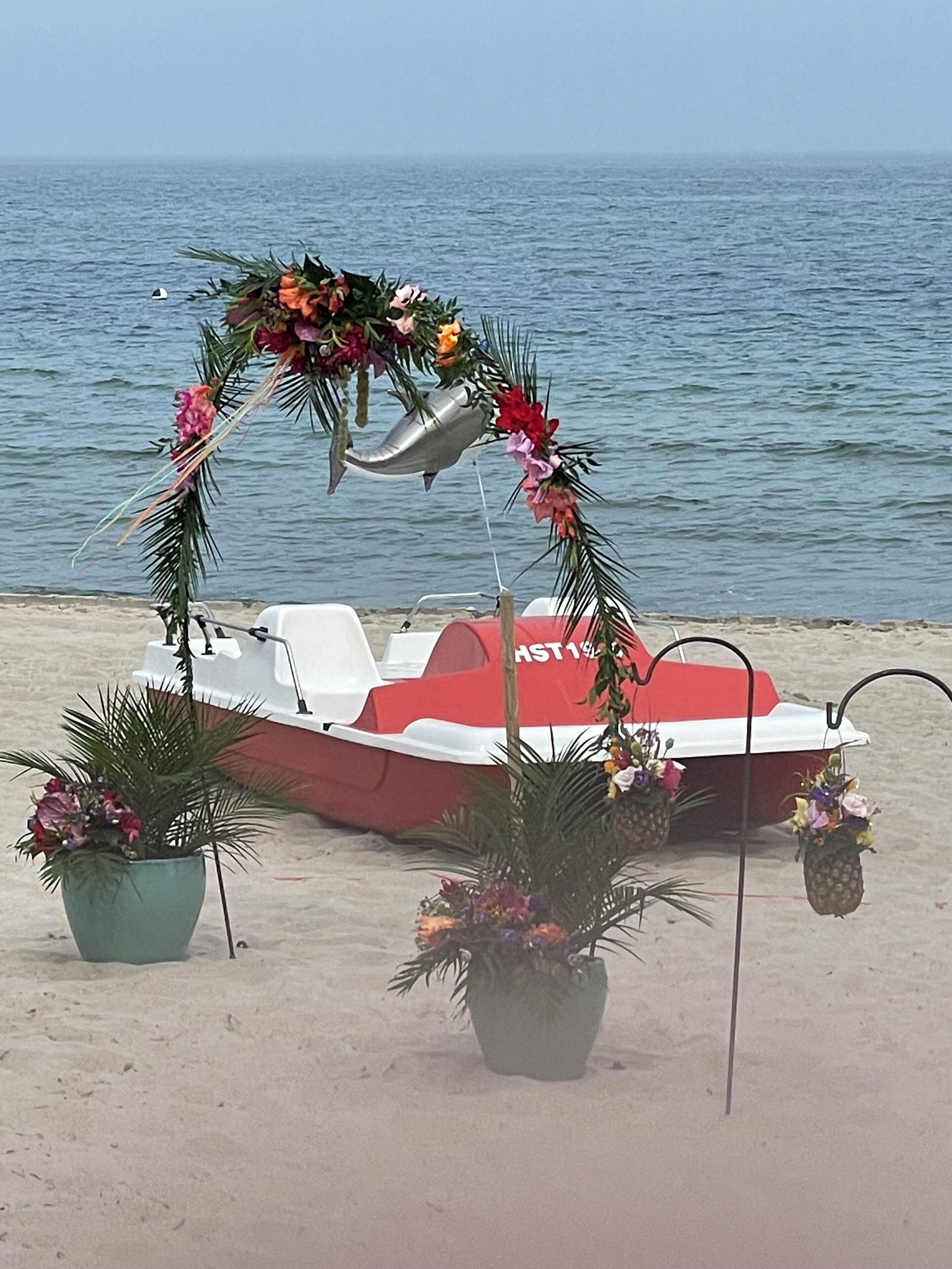 A boat decorated with flowers and a shark ornament on a beach with a calm sea in the background.