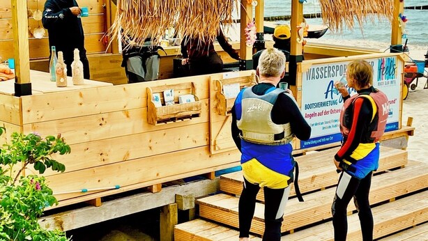 Een houten strandconstructie met een naambord, een uitkijkpost van de strandwacht en een bord van een beachclub hotel.