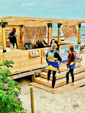 Une structure en bois installée sur la plage, avec un panneau indicateur, une tour de surveillance pour les sauveteurs et une enseigne d’hôtel faisant office de club de plage.