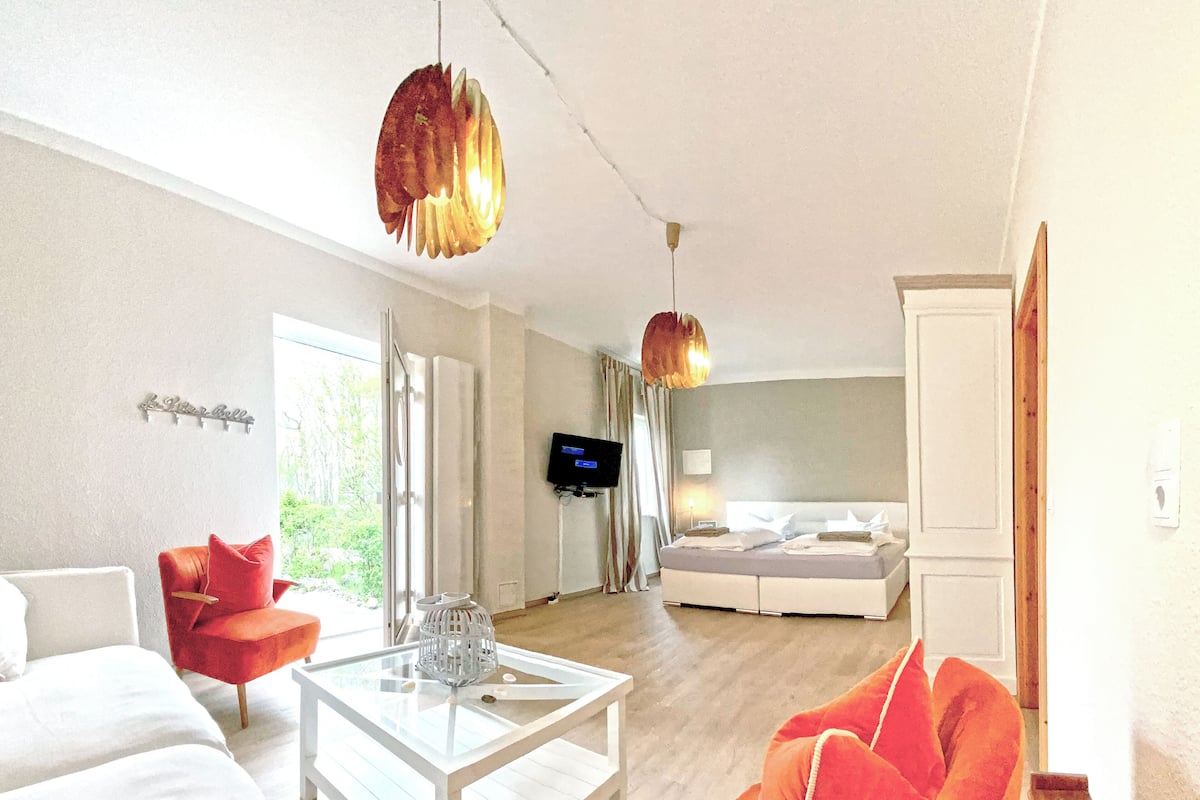 Een moderne hotelkamer met een wit bed, een rode fauteuil en een glazen salontafel. Er is een televisie aan de muur bevestigd en een groot raam zorgt voor natuurlijk licht.
