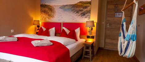 A hotel room with a bed, red bedding, a beach-themed wall mural, a hanging chair, and a wooden door.