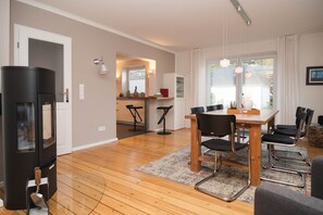 Ein modernes Wohnzimmer mit Holzboden, einem Essbereich mit Holztisch und -stühlen, einer schwarzen Unterhaltungsecke und einer Küche im Hintergrund.