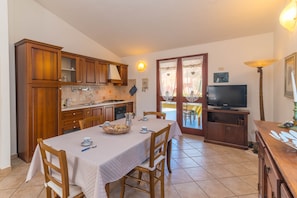 Une cuisine avec des meubles de rangement en bois, une table à manger avec des chaises, un téléviseur posé sur un meuble et une lampe.