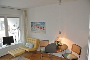 A living room with a sofa, ottoman, and two chairs around a table with a candle. A painting is on the wall, and a TV is on a stand by the window. - This small, cozy one-room apartment in Haus Brigitte... (Hörnum)