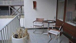 Uma pequena área de pátio ao ar livre com mesa e duas cadeiras, um vaso de plantas e um muro de tijolos brancos.