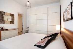 Une chambre à coucher comprenant un lit, une commode, un miroir, un luminaire au design géométrique et un mur orné d’un motif en quadrillage.