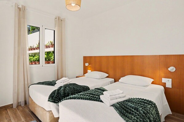 A hotel room with two beds, a wooden headboard, a window with curtains, and a view of greenery outside.