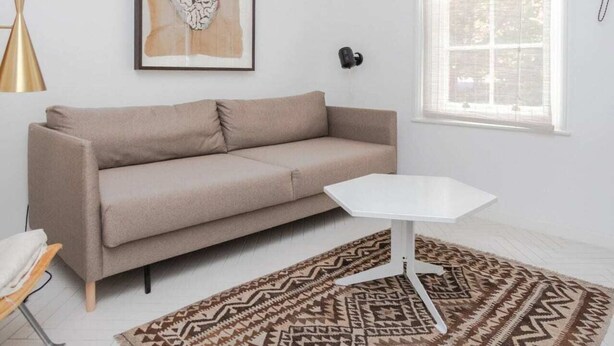 Un salon moderne avec un canapé beige, une table basse blanche, un tapis à motifs, une œuvre d’art encadrée et une fenêtre laissant entrer la lumière naturelle.