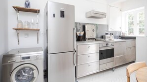 Une cuisine compacte équipée d’un réfrigérateur en acier inoxydable, d’un four encastré et d’un lave-linge. On y trouve des étagères en bois où sont disposés des bocaux et des verres, ainsi qu’une fenêtre qui laisse entrer la lumière naturelle.