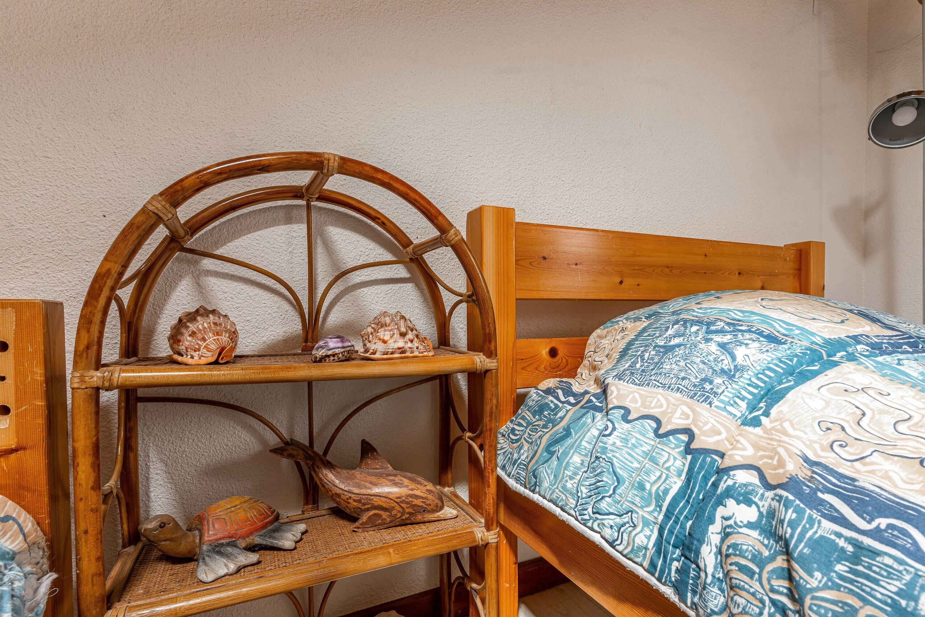 Un lit en bois recouvert d’une couverture à motifs, une chaise en bois avec une étagère au-dessus, sur laquelle sont disposés des objets décoratifs, et une lampe fixée au mur.