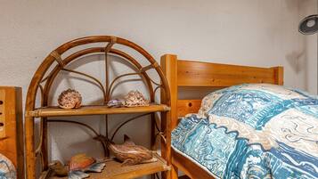 Een houten bed met een geborduurde deken, een houten stoel met een plank erboven waarop decoratieve objecten staan, en een wandlamp.