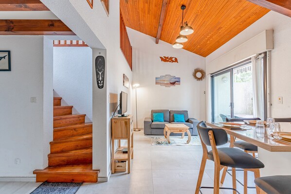 A modern interior with a wooden staircase, a dining area with a table and chairs, a sofa, and a large window.