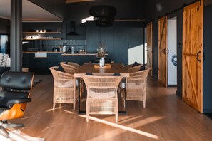 A modern dining area with a wooden table and wicker chairs, a kitchen in the background, and a large wooden door.