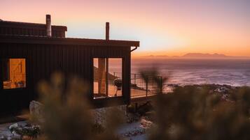 A coastal house with a wooden exterior, a stone foundation, and a view of the ocean at sunset.