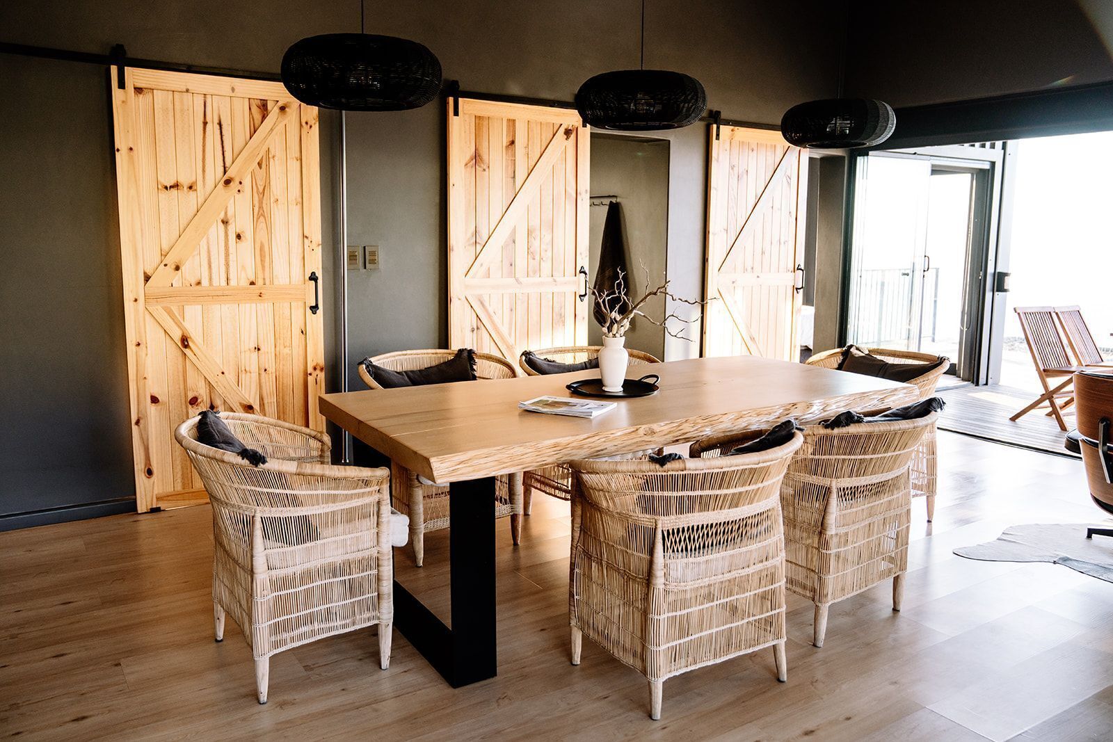 A dining area with a wooden table and wicker chairs, large wooden doors, and pendant lights.