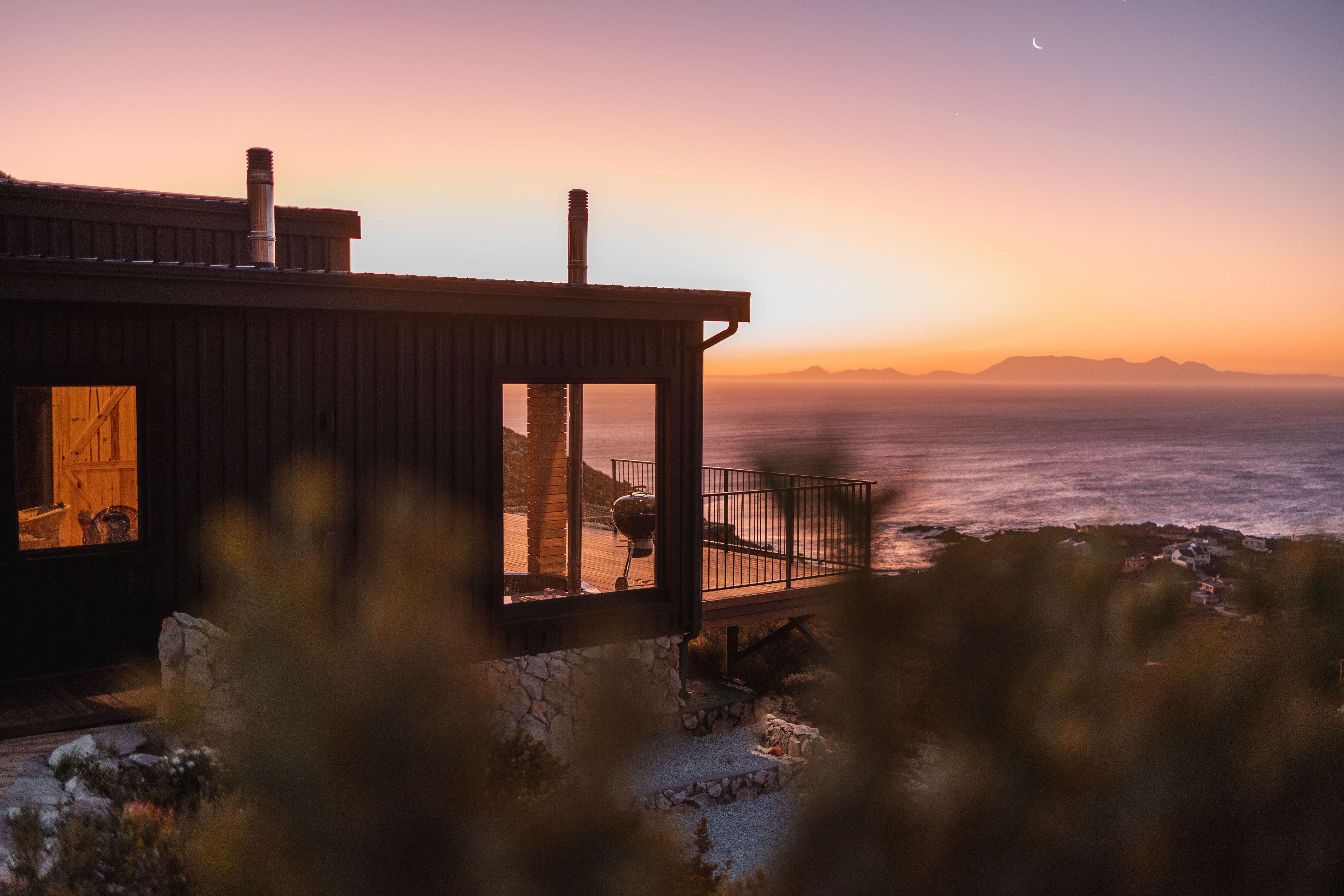 A coastal house with a wooden exterior, a stone foundation, and a view of the ocean at sunset.