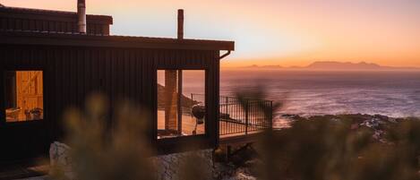 A coastal house with a wooden exterior, a stone foundation, and a view of the ocean at sunset.