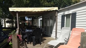 Un patio couvert avec une table et des chaises, un barbecue, et une maison mobile blanche dotée de volets.