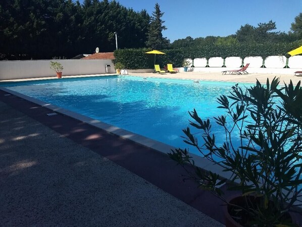 Une piscine à l’eau d’un bleu cristallin, entourée d’une terrasse dallée. On aperçoit, au fond, des parasols jaunes et des chaises longues.