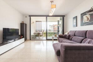 Een moderne woonkamer met een flatscreen televisie, een bank, een eethoek met stoelen en een schuifpui naar een balkon.